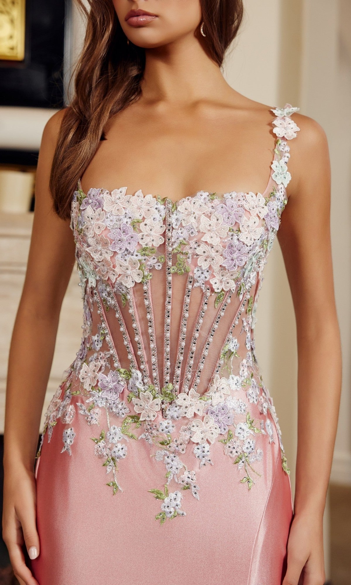 A woman wears the Nox Anabel M1755 long prom dress with a sleeveless sheer structured bodice, 3D floral appliques, and a flowing floor-length satin skirt in soft pink.
